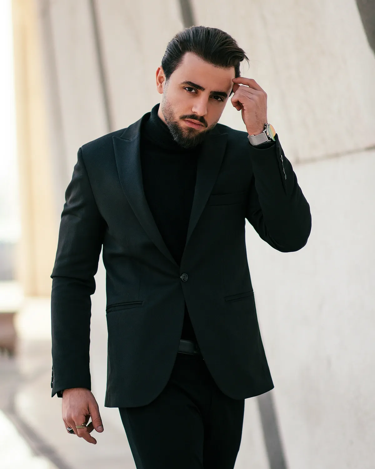 Stylish man in a black suit and turtleneck walks confidently, adjusting his hair against a modern architectural backdrop.